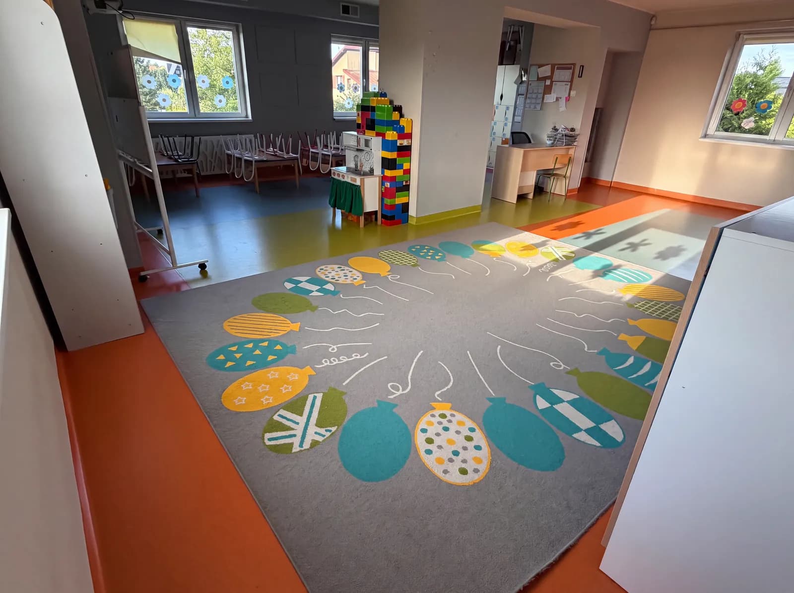 Sala przedszkolna z kolorową podłogą, zabawkową kuchnią i wieżą z dużych klocków